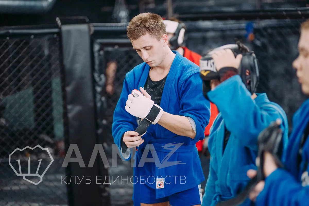 Боец в синей самбовке надевает перчатку перед тренировкой, рядом готовятся остальные