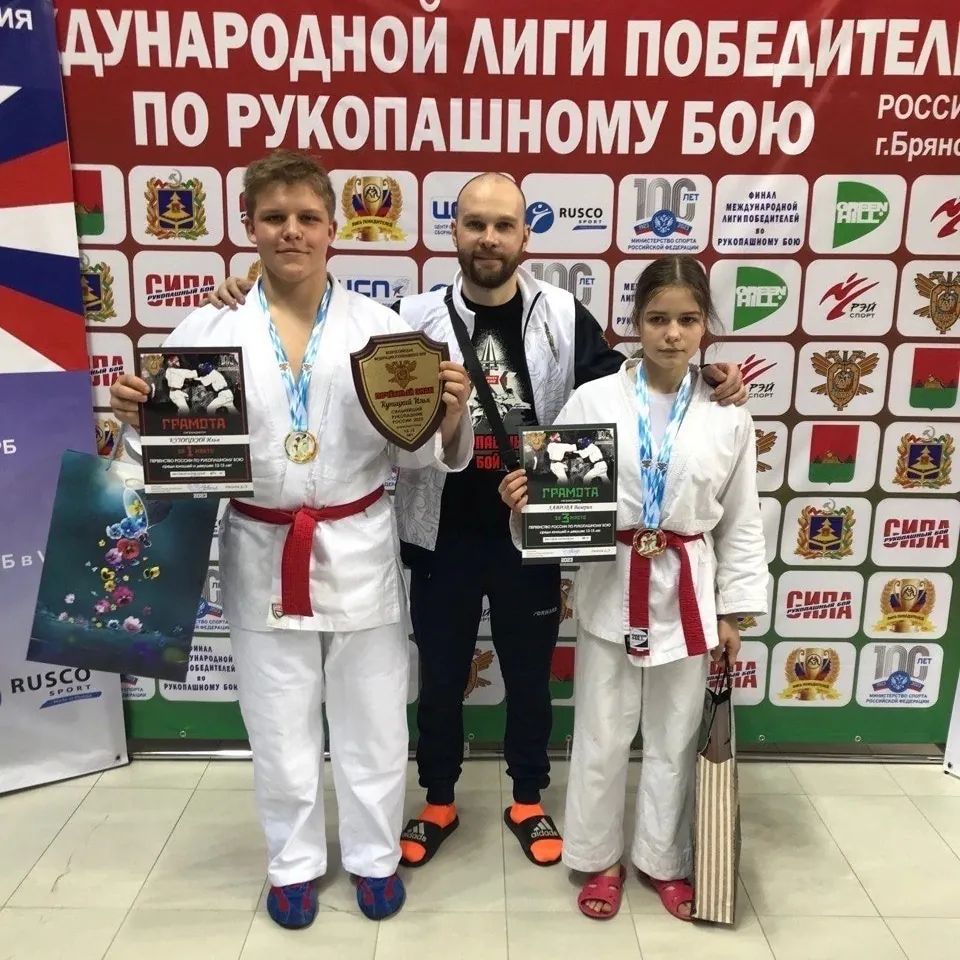 Подросток и девочка в белых кимоно с медалями и грамотами, между ними тренер; у подростка в руках наградной щит, за ними баннер турнира по рукопашному бою