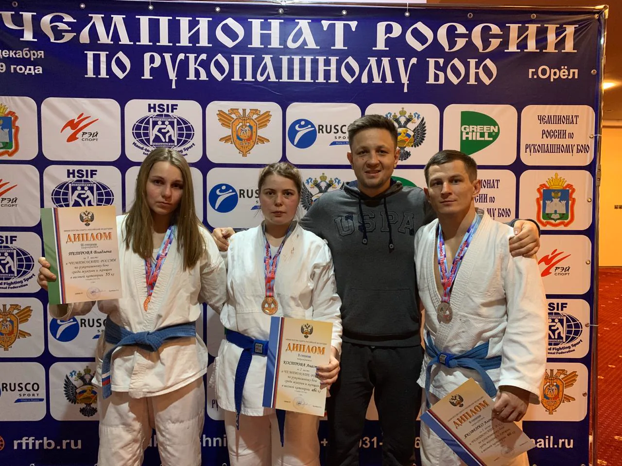 Три спортсмена клуба в белых кимоно с медалями и дипломами и тренер на фоне баннера «Чемпионат России по рукопашному бою, г. Орёл»
